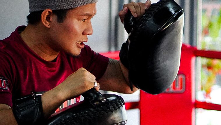 Head trainer Kru Maew in a maroon KRU shirt and beanie, holding focus pads in the ring